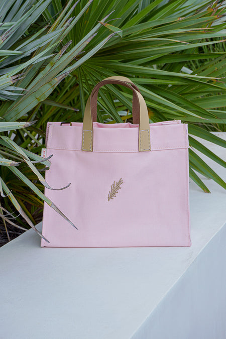 Pink tote bag with beige handles on a light surface with green foliage in the background